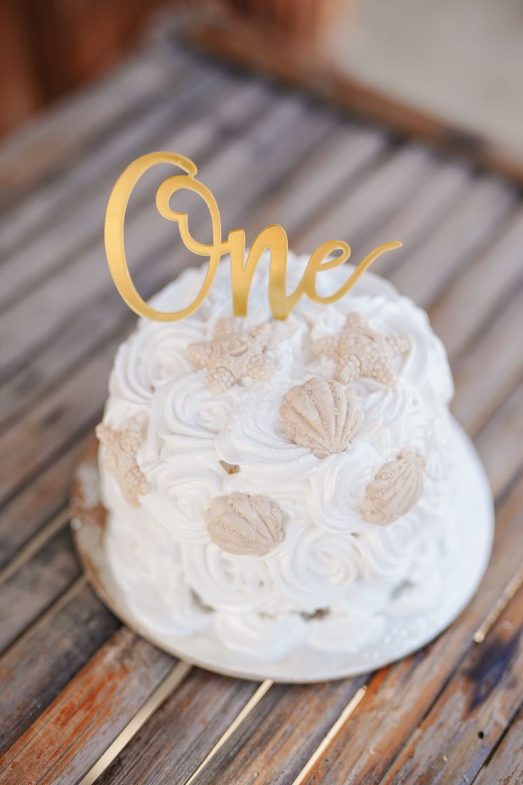 a white cake with gold lettering on top sitting on a wooden table next to a cup