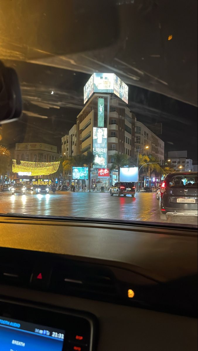 the view from inside a car driving down a city street at night with buildings lit up