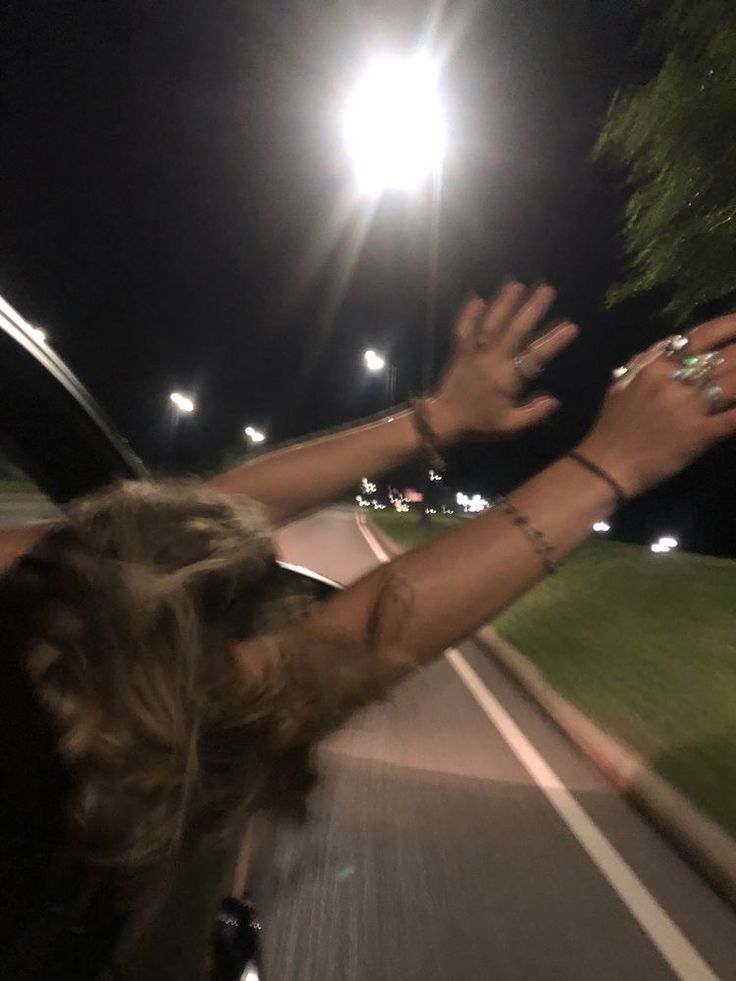 two people holding their hands up in the air as they drive down a street at night