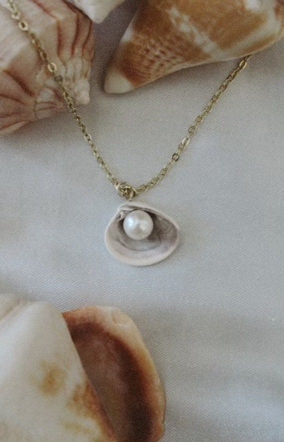 Our Pearl Seashell Necklace is such a fun addition to your summer jewelry collection! This necklace features a real seashell with a white pearl inside, strung on a classic gold chain.  This necklace is handmade with real seashells. You can customize and choose from our shell options including:  -Shell from Florida  -Shell from Outerbanks  -Shell from Chincoteague Beach  Inspired by the coast, this necklace would be perfect to wear in the summertime or on your next seaside vacation! All of our jewelry is handmade, each piece beautiful and unique. Pearl Pendant Shell Gift, Pearl White Shell With Pearl Charm, Pearl Pendant Shell As Gift, Pearl Shell With Pearl Charm, Ocean-inspired Shell Necklace With Pearl Charm As Gift, Ocean-inspired Shell Necklace With Pearl Charm For Gifts, White Shell With Pearl Pendant, Pearl Drop Shell, Gift Pearl Drop Shell Made Of Mother Of Pearl