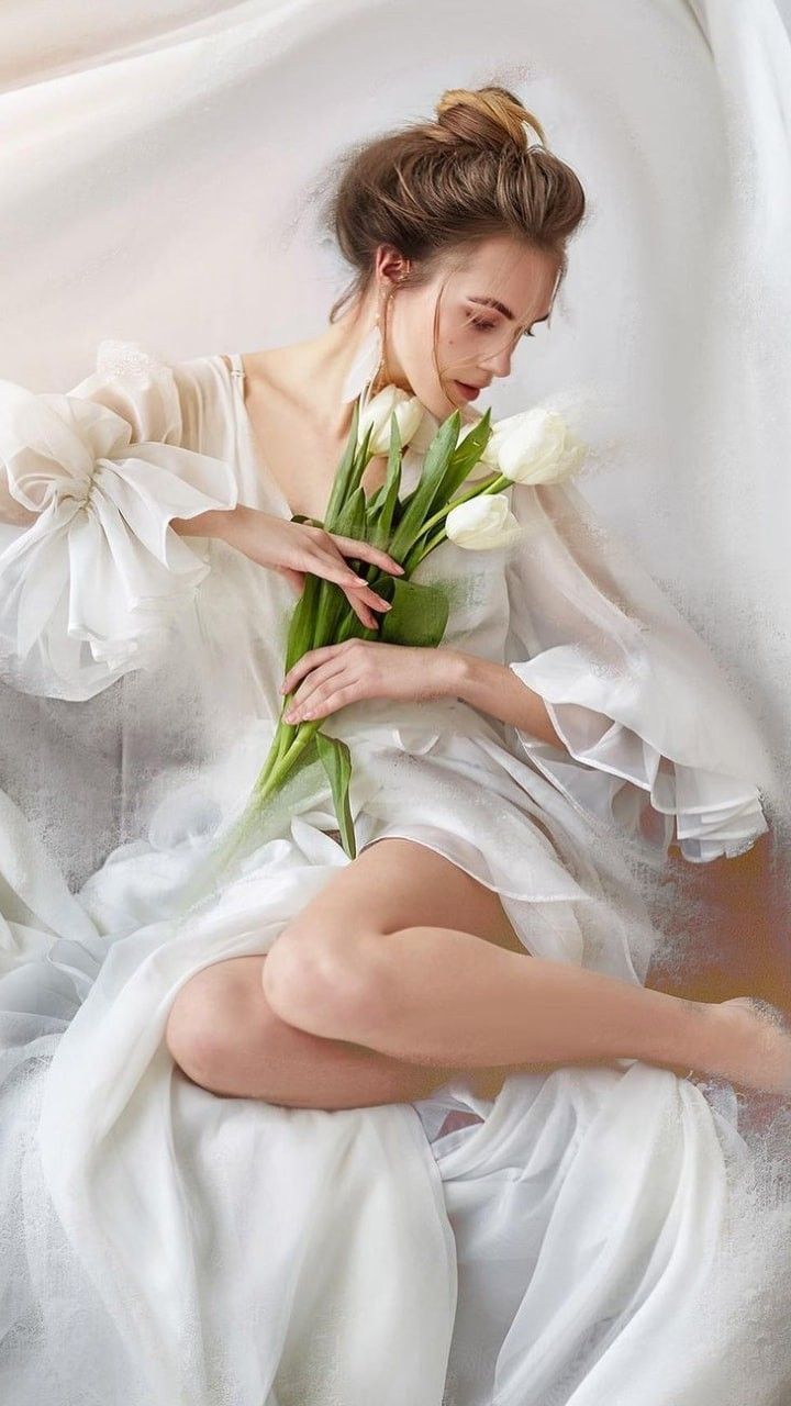 a woman in a white dress with flowers on her lap, sitting down and looking at the camera