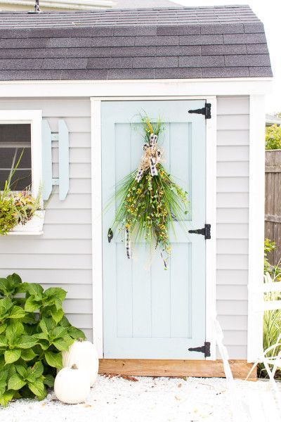 a white shed with a wreath on the door and some green plants in front of it