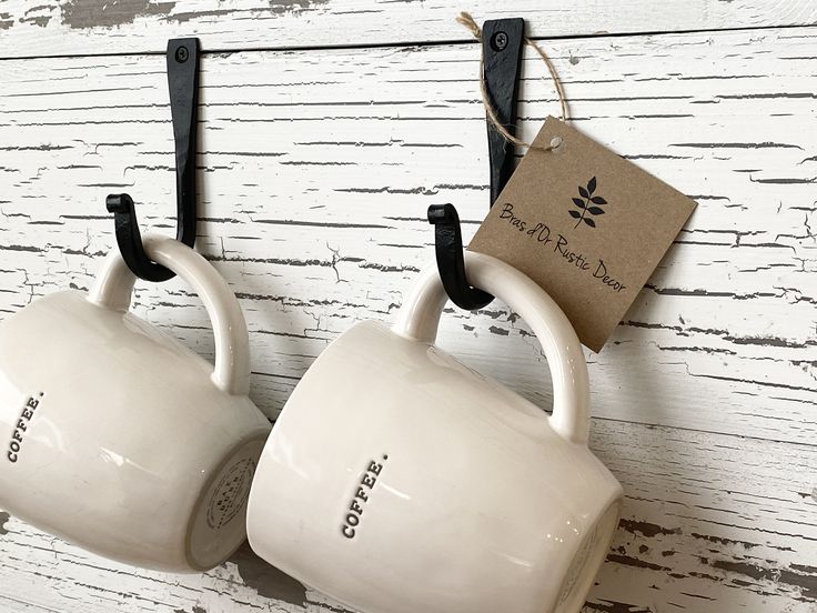 two coffee mugs hanging from hooks on a white wooden wall with a tag attached to them