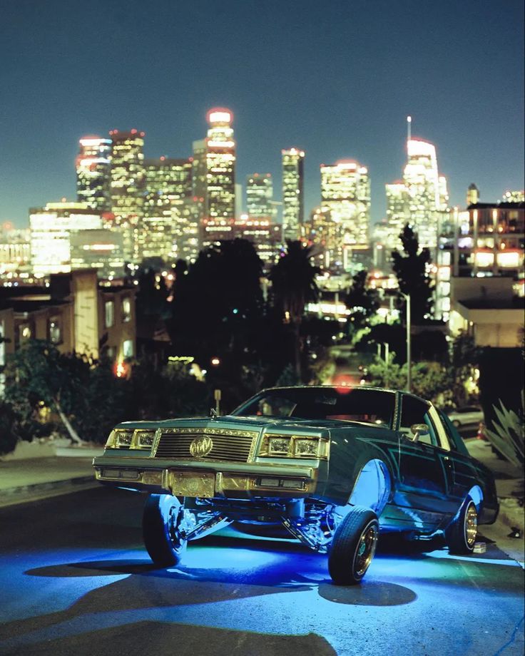 an old car is parked in front of a city skyline at night with lights on