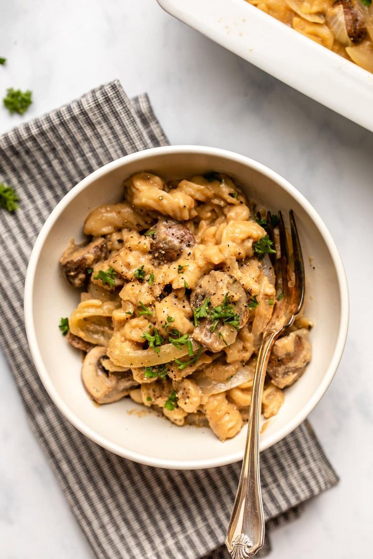 a white bowl filled with pasta and mushrooms next to a dish on a gray napkin