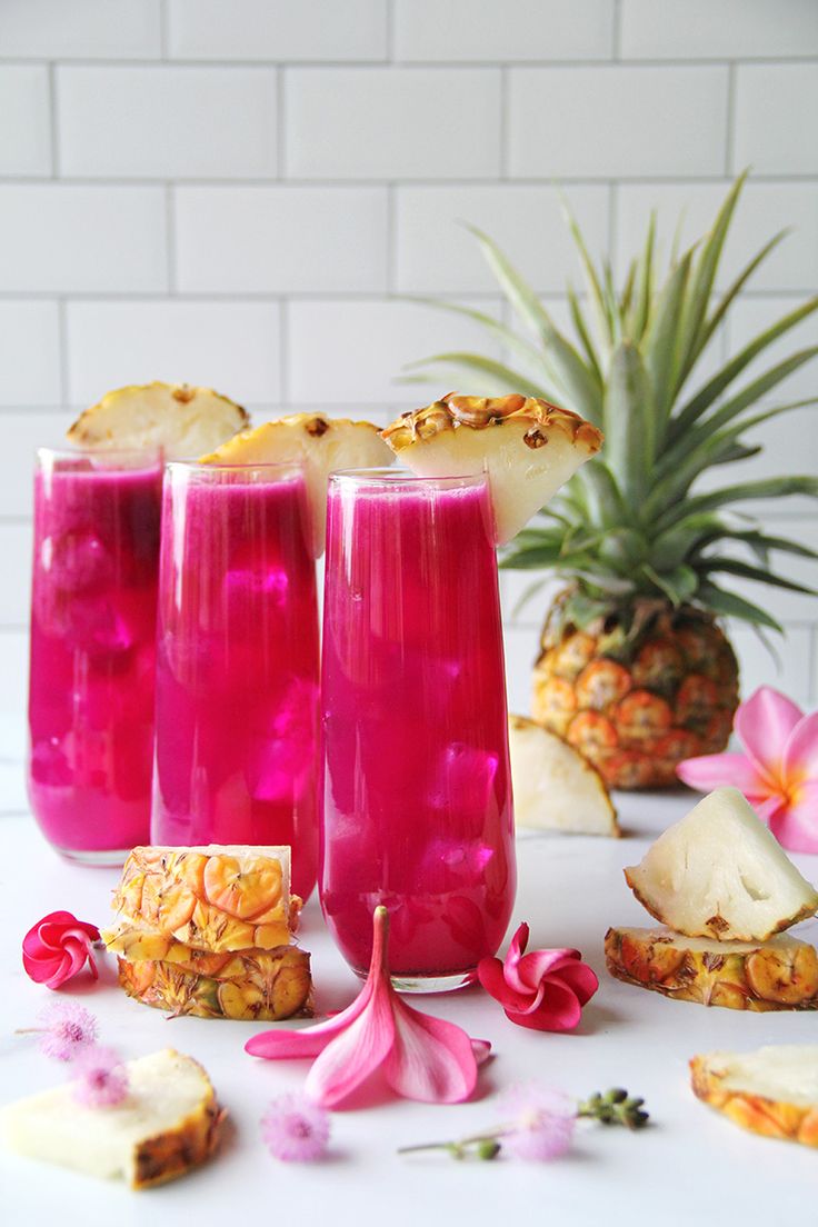 three glasses filled with pink liquid next to sliced pineapples and other food items