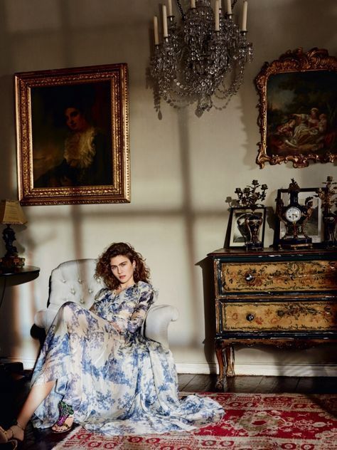 a woman sitting on a chair in a room with paintings and chandelier above her