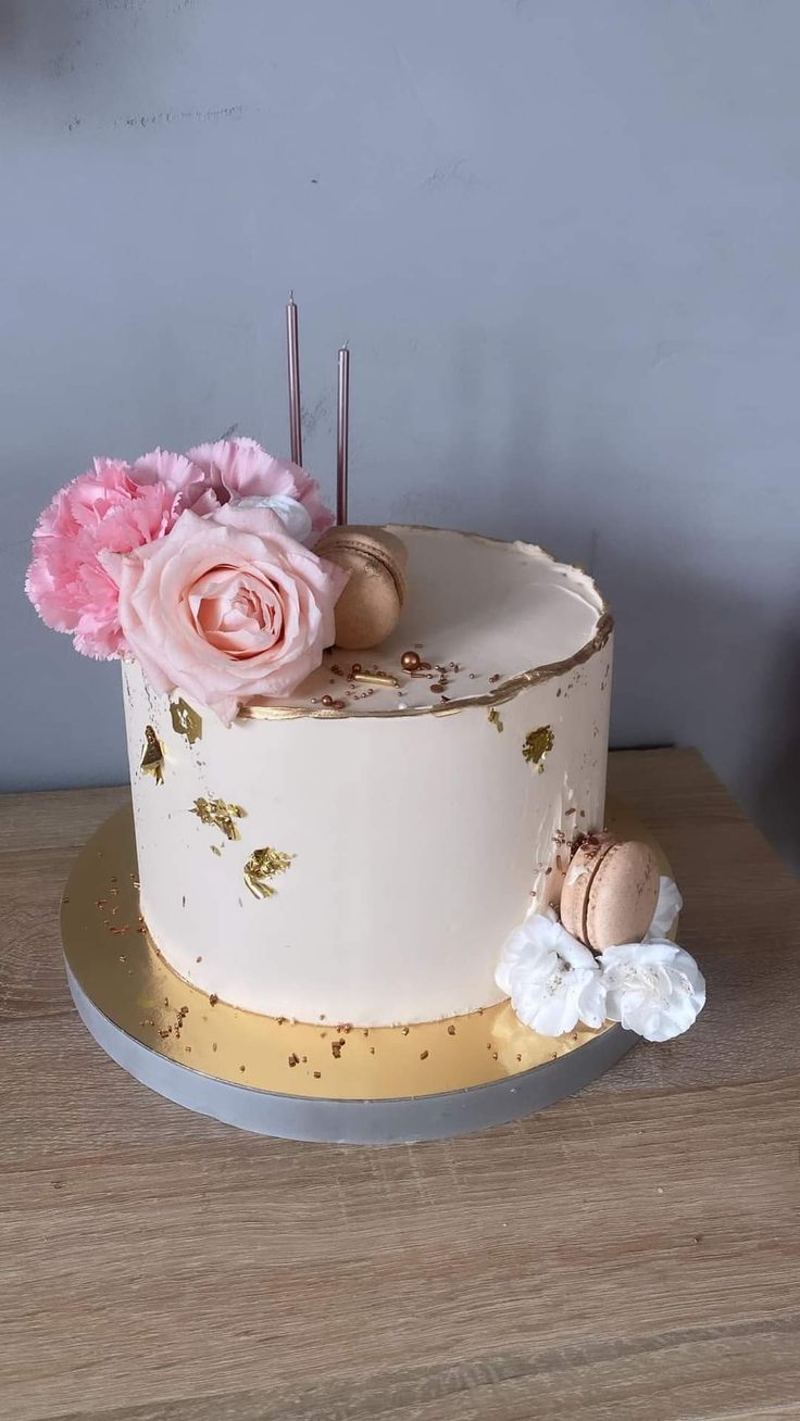 a white cake with pink flowers on top sitting on a wooden table in front of a gray wall