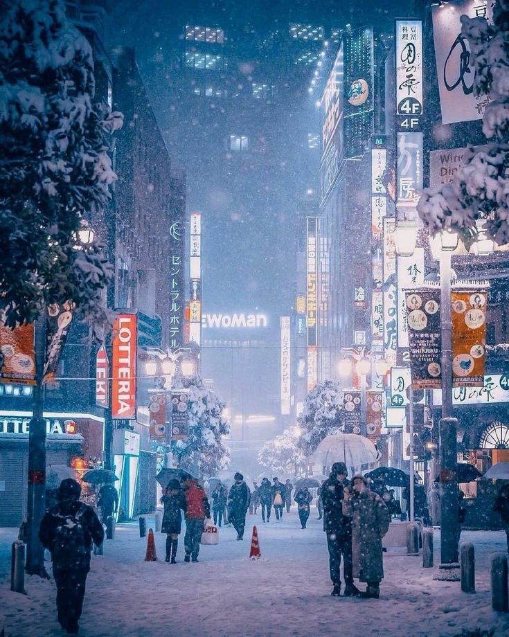 people walking in the snow on a city street at night with lights and signs all around