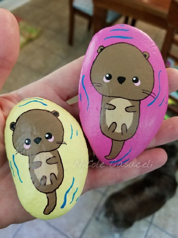 two painted rocks in the shape of otters are being held by someone's hand