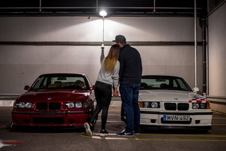 a man and woman standing next to two parked cars in a parking lot at night