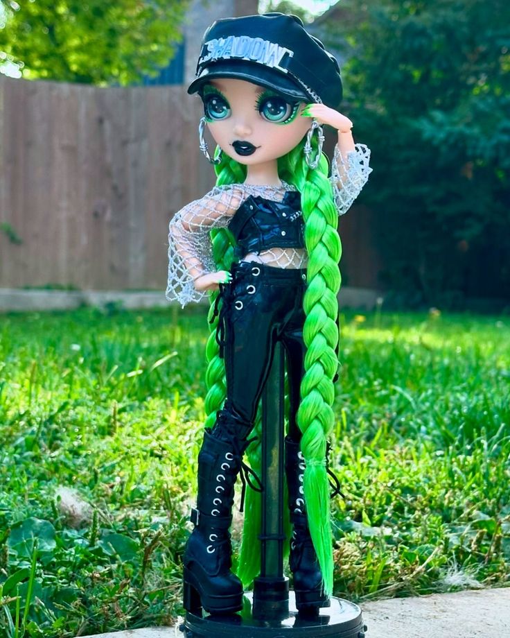 a doll with long green hair and black pants standing in the grass next to a fence