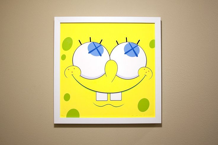 a yellow and green square with two blue eyes on it's face in front of a beige wall