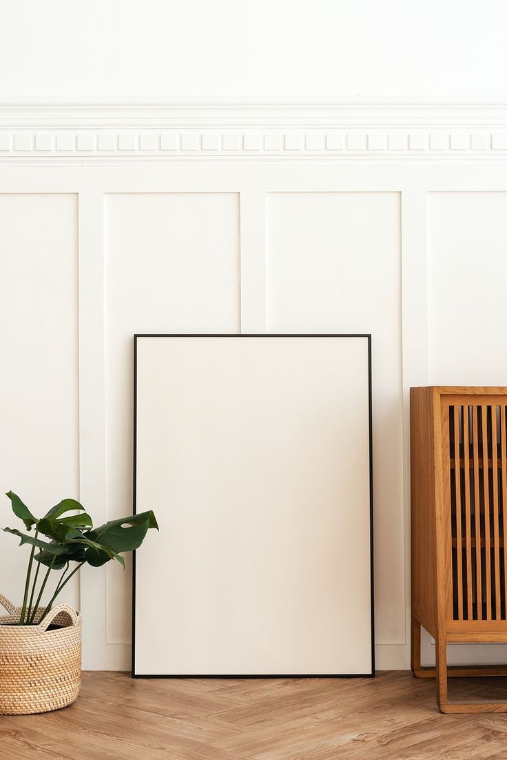 an empty frame next to a plant in a room with white walls and wood flooring