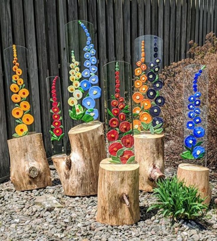 colorful glass flowers are displayed on wooden stumps in a garden area with gravel and rocks