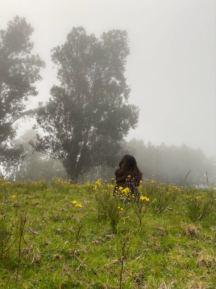 a woman sitting on top of a lush green field next to a forest filled with lots of trees