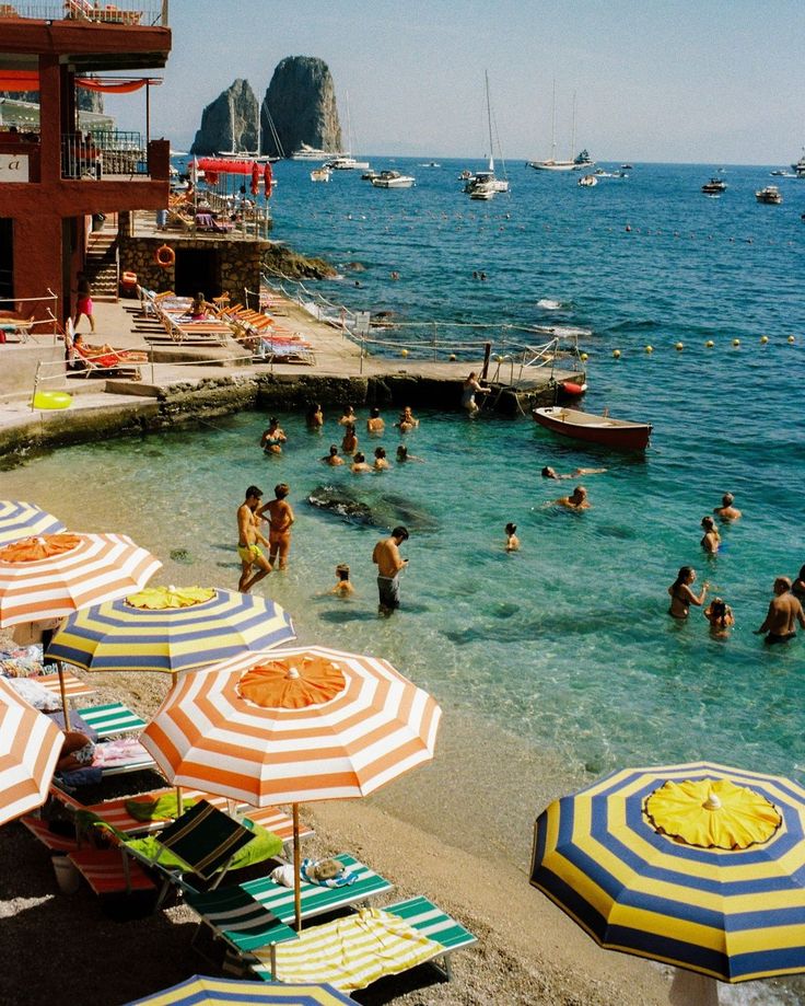 many people are on the beach with umbrellas and boats in the water behind them