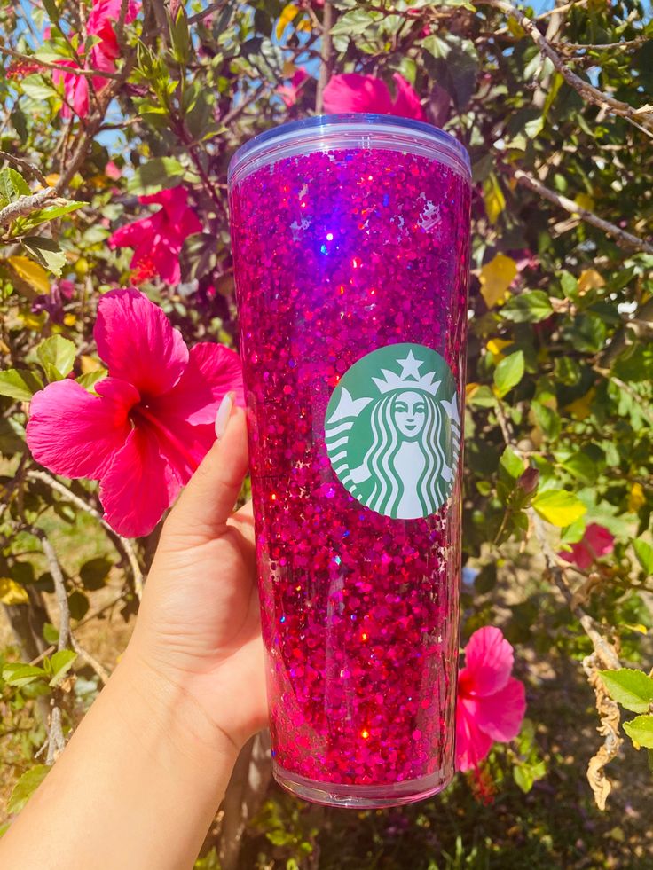 a hand holding up a pink starbucks cup with purple glitter on it and flowers in the background