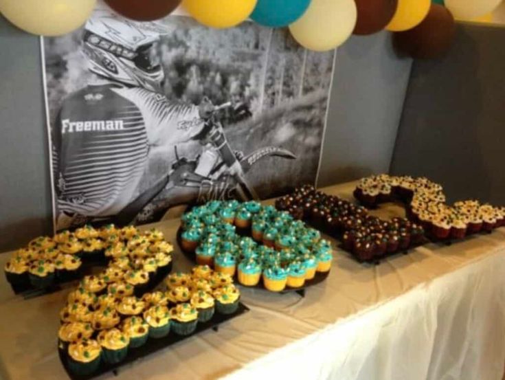 a table topped with lots of cupcakes covered in frosting next to balloons