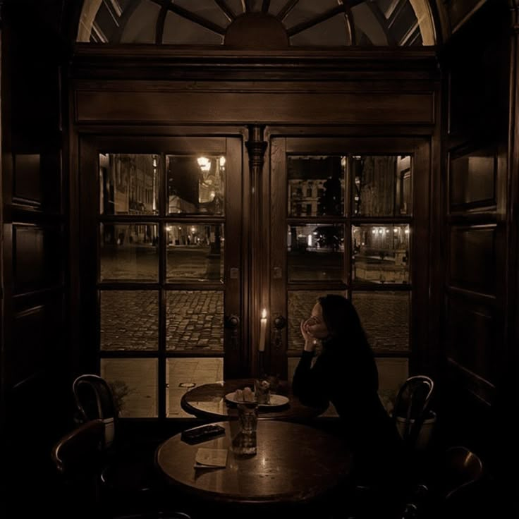 a woman sitting at a table with a candle in her hand and looking out the window