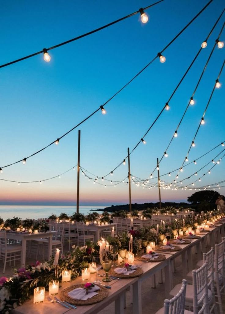 a long table is set up with candles and flowers for an outdoor dinner by the ocean