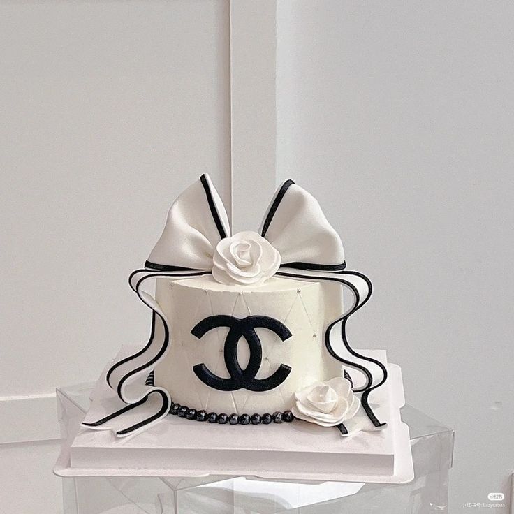 a chanel cake with white flowers and black ribbon on top is sitting on a clear plate