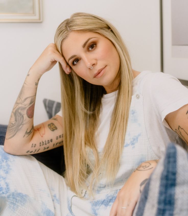 a woman with long blonde hair and tattoos on her arms, sitting in a chair