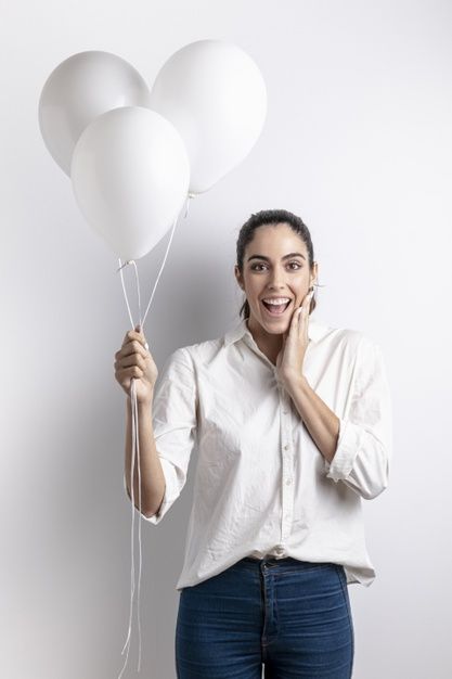 a woman holding three white balloons in one hand and smiling at the camera while standing against a wall