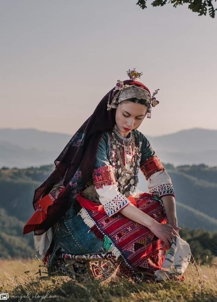 a woman sitting on top of a grass covered field next to a tree and mountains