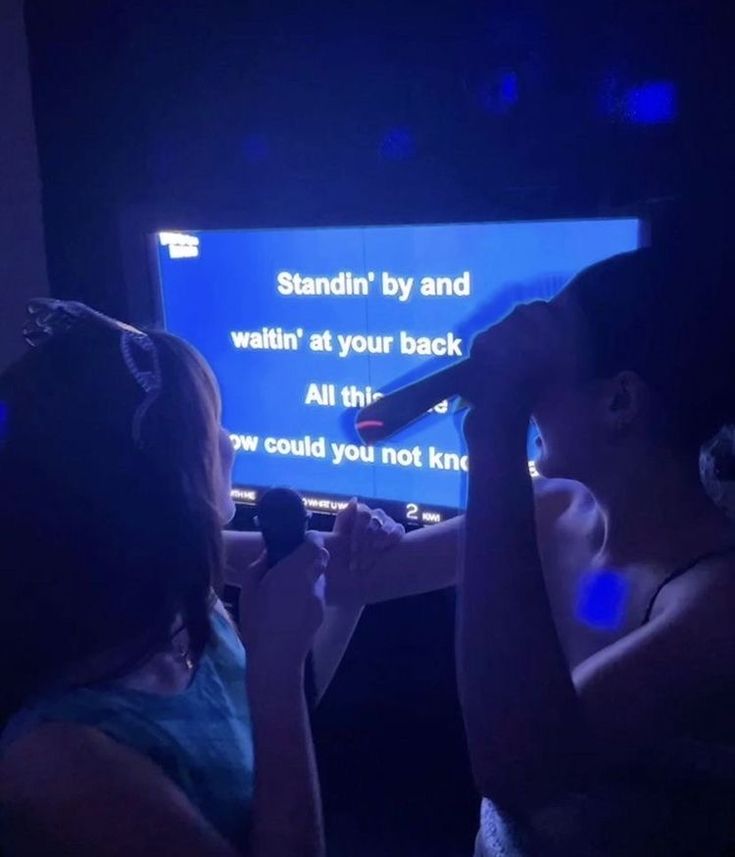 two girls are watching television in the dark with their hands on their hipss and one is holding a microphone