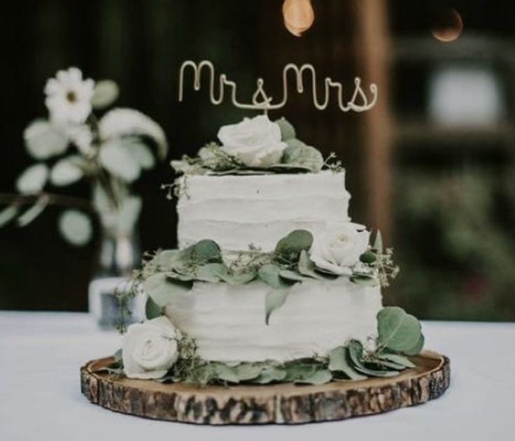 a white wedding cake with greenery and the word mr and mrs on top sitting on a table