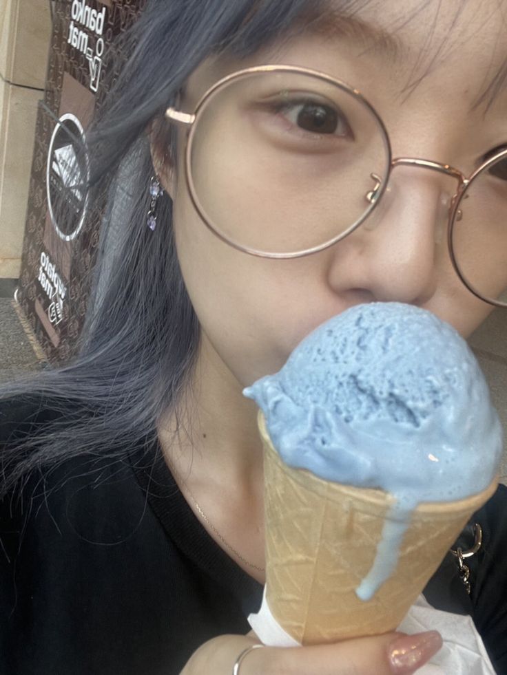 a woman wearing glasses is eating an ice cream cone with blue icing on it