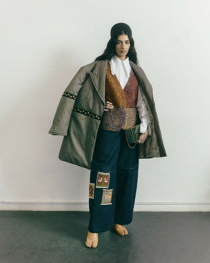 a woman standing in front of a white wall wearing a coat and pants with patches on it