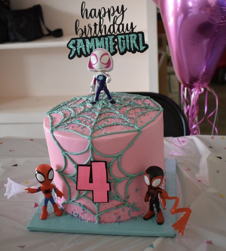 a pink birthday cake with spiderman figurines on top and balloons in the background