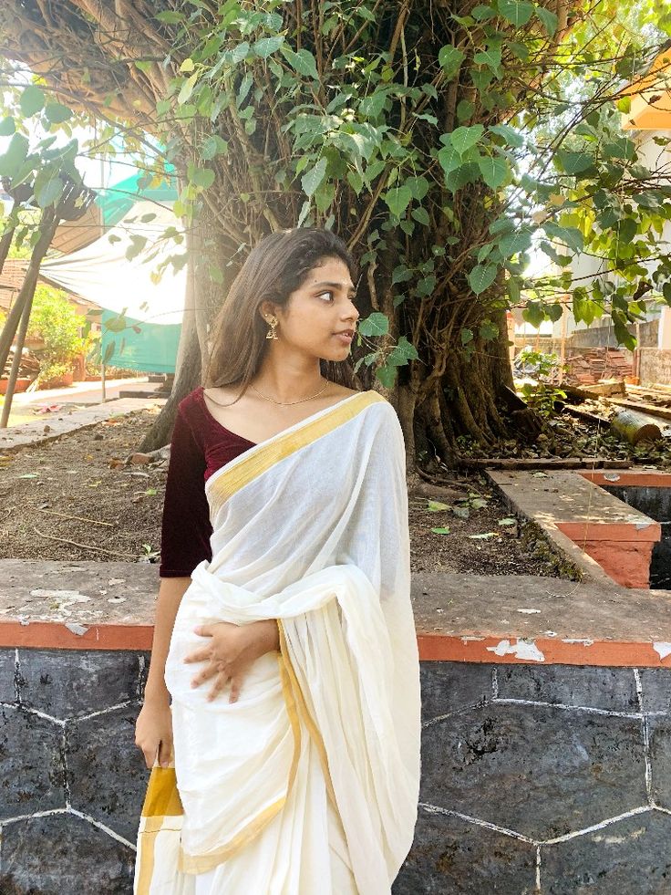 a woman standing in front of a tree wearing a white and yellow sari with gold border