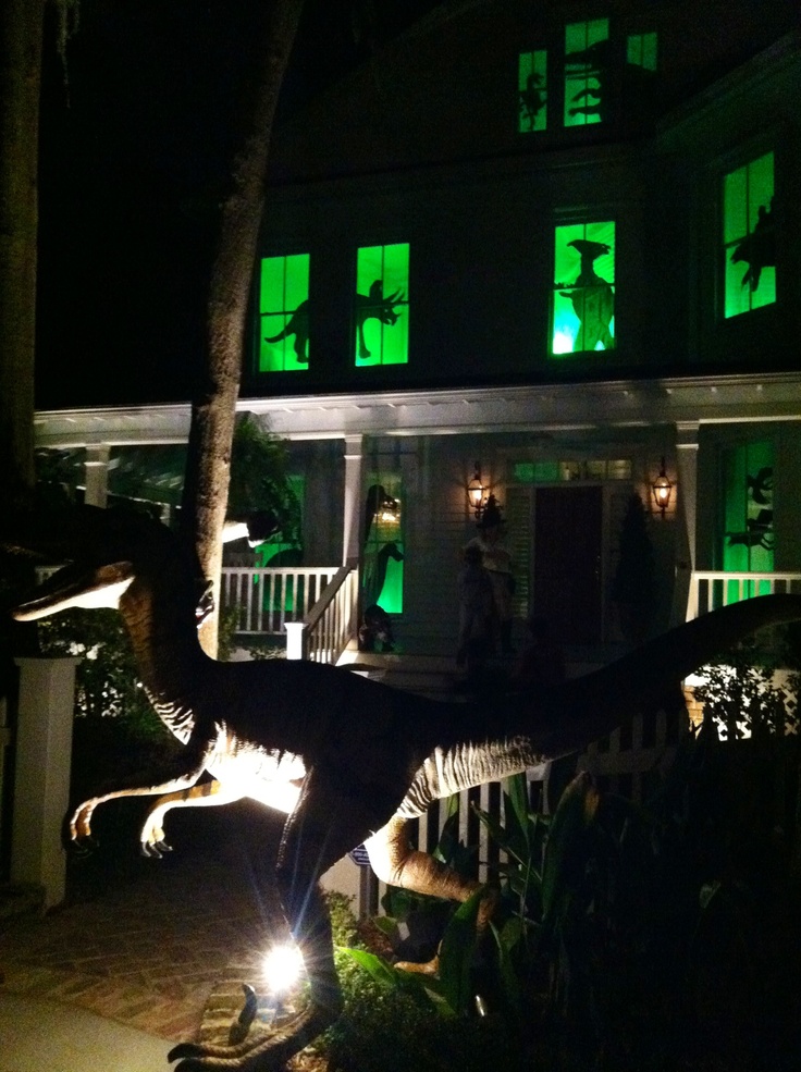 a dinosaur statue in front of a house with green lights on it's windows