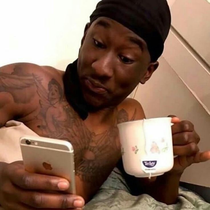 a shirtless man holding a cup and cell phone in his hand while sitting on a bed