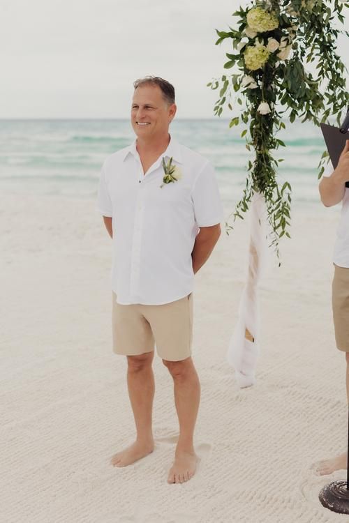 two men are standing on the beach under an arch decorated with flowers and greenery