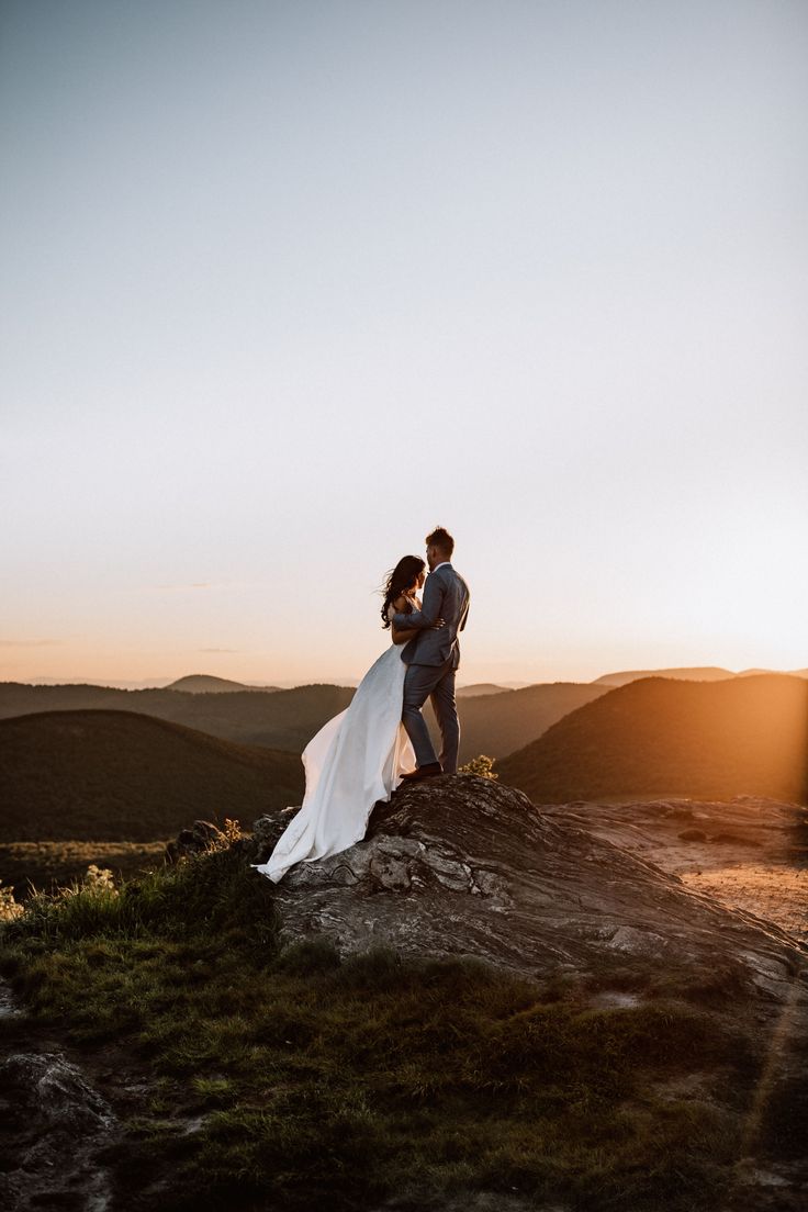 a bride and groom standing on top of a mountain at sunset with the sun behind them