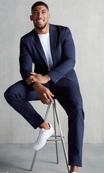 a man in a suit sitting on a stool with his legs crossed, smiling at the camera