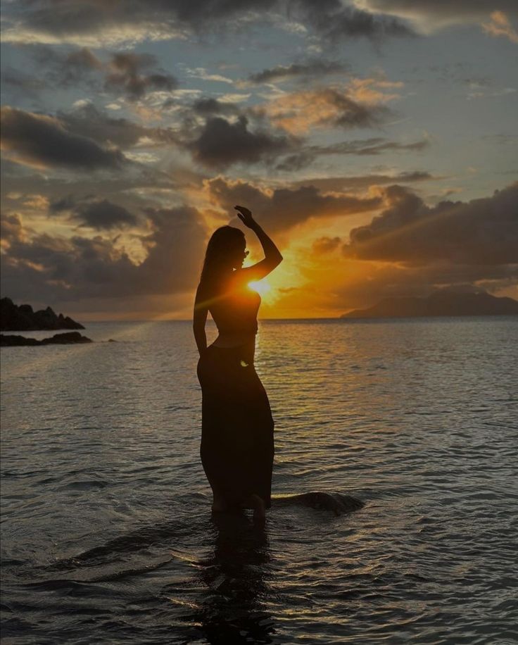 a woman standing in the water at sunset with her arms up and hands behind her head