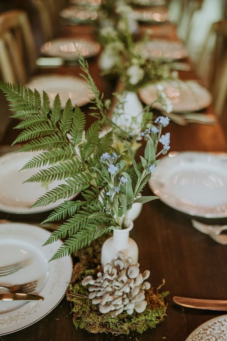 the table is set with white plates, silverware and green plants in vases