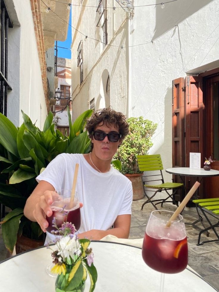 a man sitting at an outdoor table with two drinks in front of him and another person holding a straw