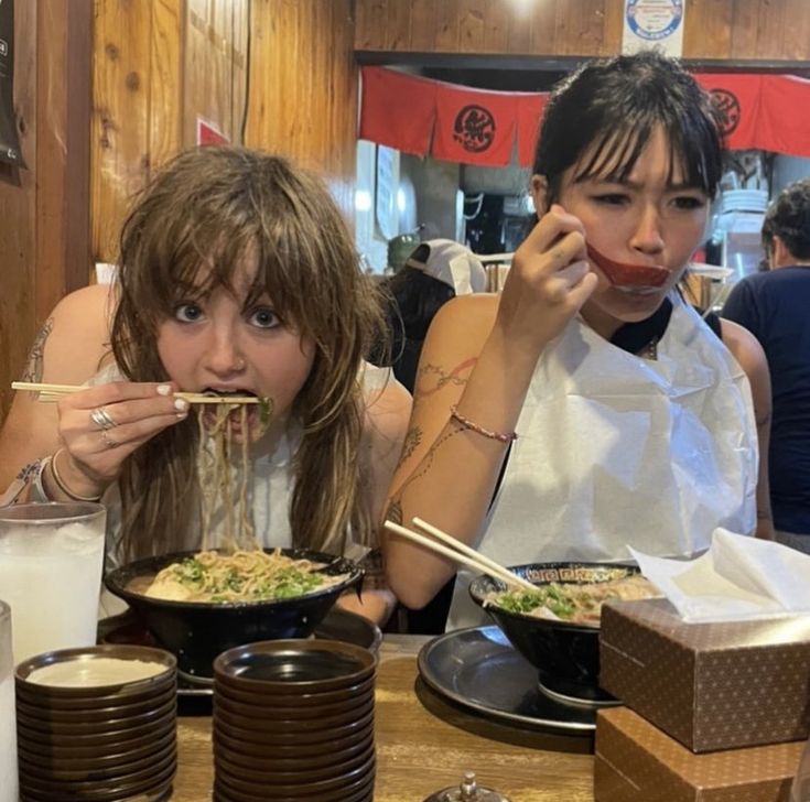 two women sitting at a table eating food with chopsticks in their mouths and holding chop sticks up to their mouth