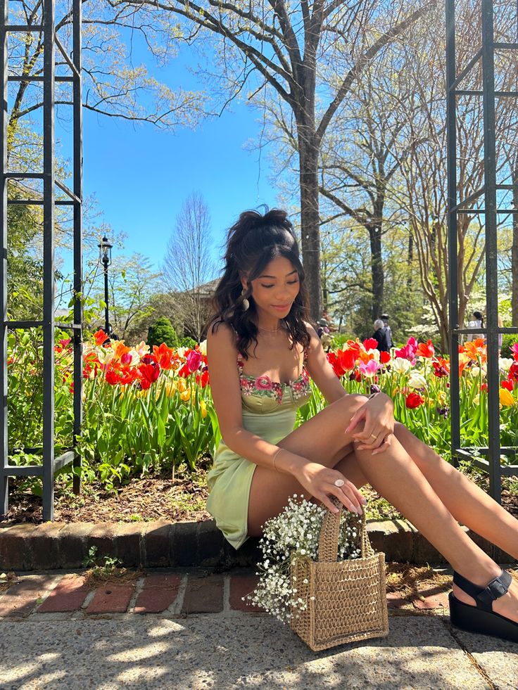 a woman sitting on the ground in front of some flowers and holding a purse with her legs