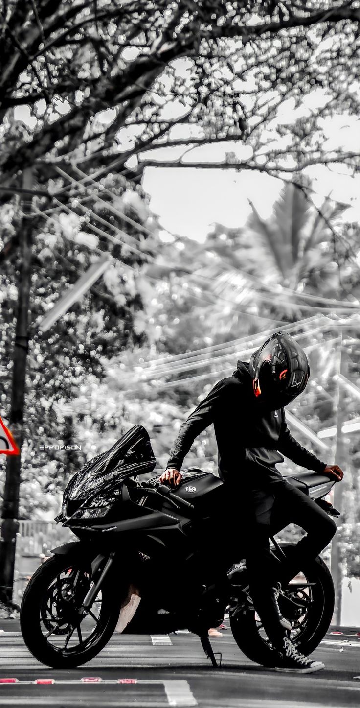 a man riding on the back of a black motorcycle down a street next to a tree