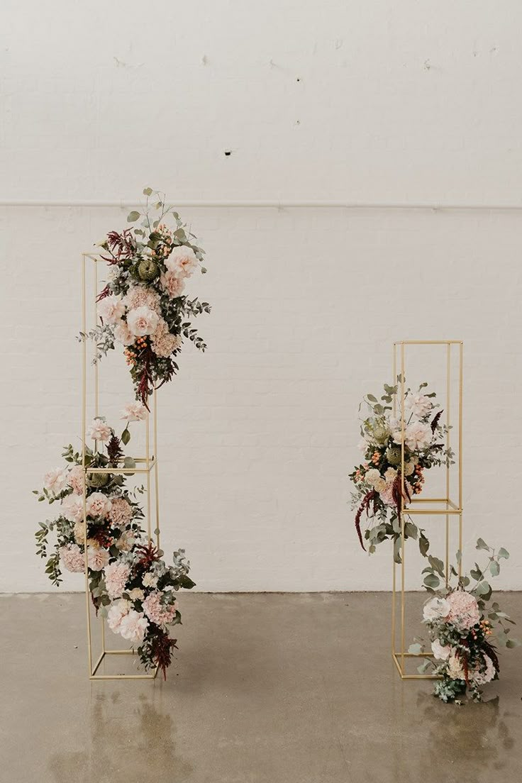 two gold stands with flowers and greenery on them in front of a white wall