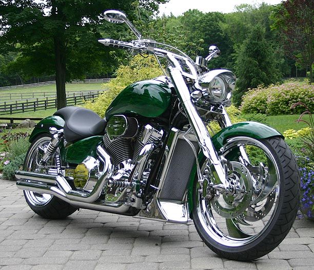 a green motorcycle parked on top of a brick road next to flowers and trees in the background