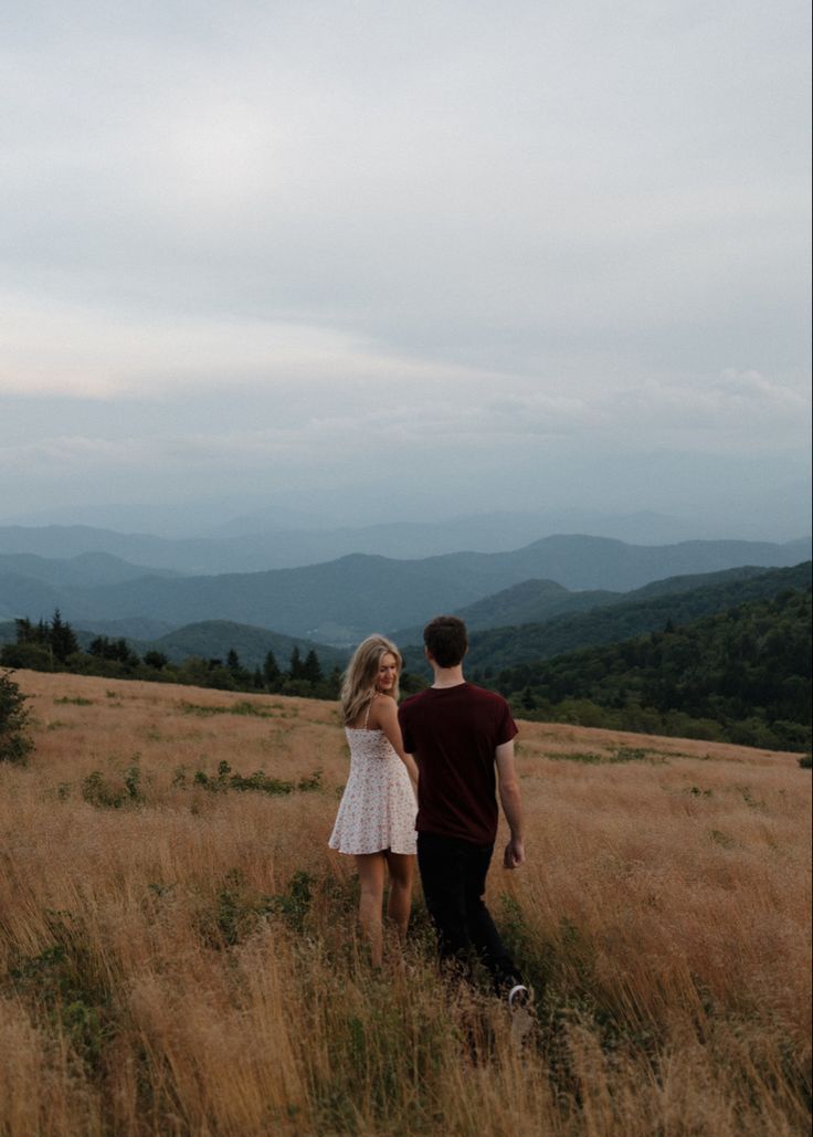 a man and woman standing in the middle of a field with mountains in the background