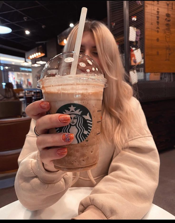 a woman sitting at a table with a starbucks drink in her hand and holding it up to the camera